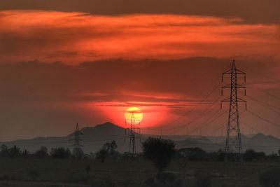Silhouette electricity pylons on landscape against orange sky