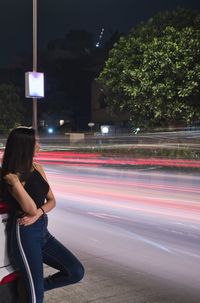 Young woman standing on sidewalk by light trails at night