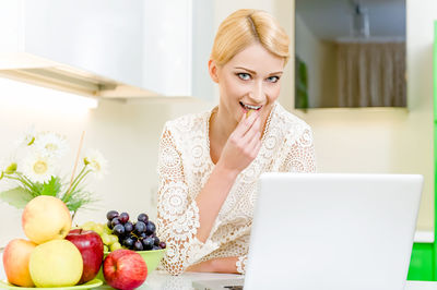 Portrait of young woman using laptop while standing by fruits on table