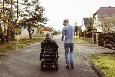 Rear view of senior man with disability in motorized wheelchair by young caregiver walking on road