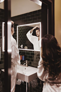 Woman standing at entrance of bathroom while man looking in mirror