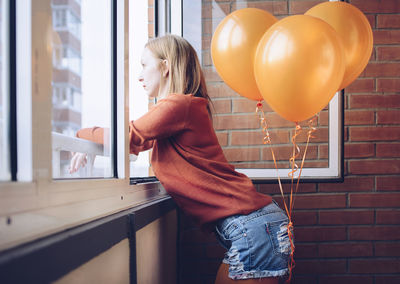 Side view of woman with orange balloons looking through window while standing at home