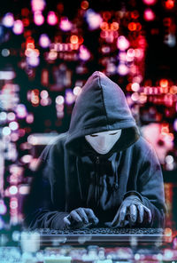 Computer hacker wearing mask and hooded shirt while tying on keyboard against illuminated lights