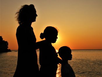 Side view of silhouette mother with daughters standing at beach against sky during sunset