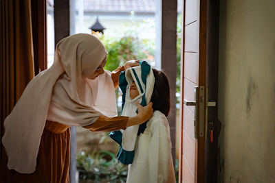 Smiling mother assisting daughter in wearing hijab