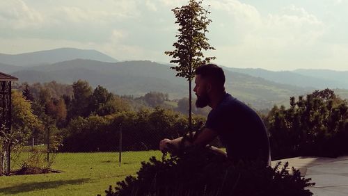 Side view of young man sitting on mountain against sky