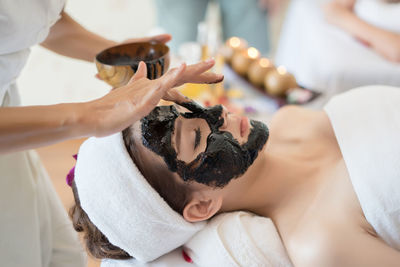 Beautician applying coal facial mask on face of young woman in spa