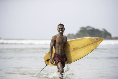 Portrait of shirtless male surfer carrying surfboard in sea