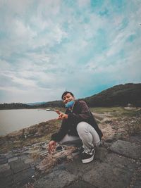 Young man sitting on rock against sky