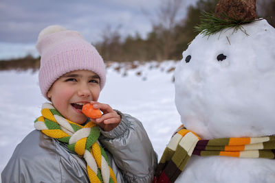 Girl in scarf bites carrot, which was the nose of snowman, smiling. winter family weekend, lifestyle