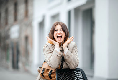 Exuberant young woman with hands on cheeks, excitedly smiling on urban chair