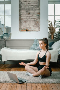 Cheerful sportswoman with blond hair meditates in lotus position, resting on the floor at home 