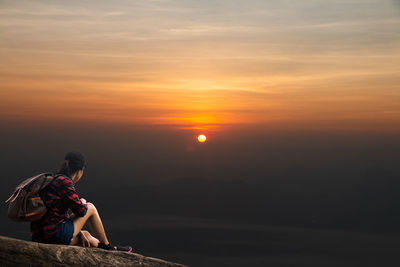Man sitting on rock against sky during sunset