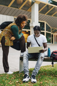 Male student e-learning on laptop while female friend standing by in campus