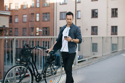 Man using mobile phone while walking with bicycle on footbridge against buildings in city