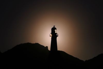 Low angle view of silhouette lighthouse by building against sky during sunset