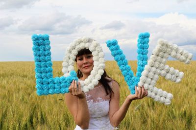 Portrait of beautiful woman holding love text while standing on grassy field against cloudy sky