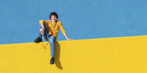 Boy listening to music through headphones while sitting on yellow railing against blue wall