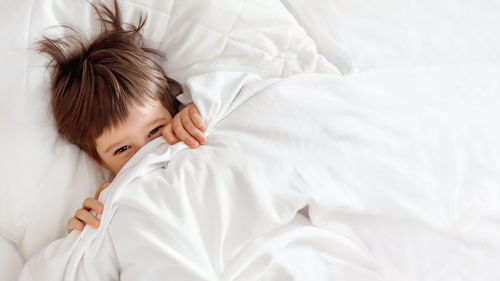 Cute toddler hides in bed. disheveled boy wakes up and peeps out from under white blanket. morning.