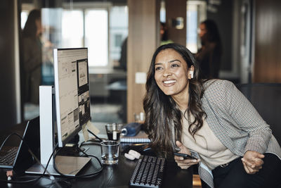 Happy businesswoman wearing wireless in-ear headphones sitting at desk in office