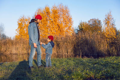 Father and son in red knitted hats are standing in autumn in the village by the lake at sunset