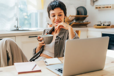 A young smiling woman sitting in the kitchen uses a laptop for work and drinks coffee.