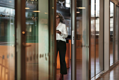 Full length of confident senior businesswoman using smart phone while reflecting on glass door at office