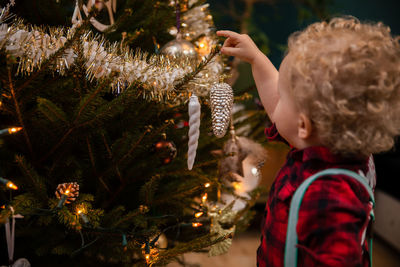 A two-year-old boy with blond curls hangs a bauble on a christmas tree.