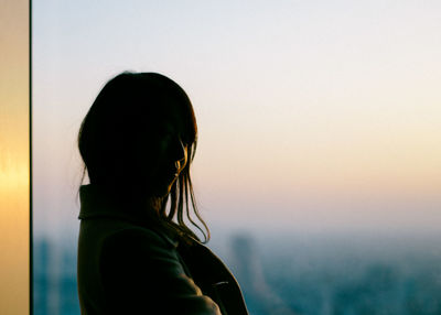 Side view of woman standing by window during sunset