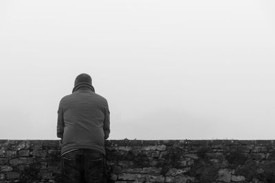 Rear view of man leaning on retaining wall against clear sky