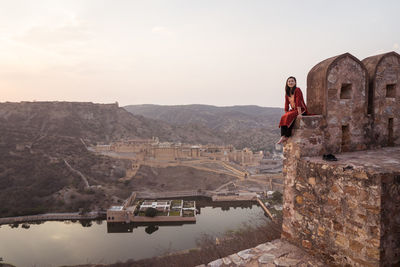 Side view of ethnic sian female tourist sitting looking at camera on aged stone wall against mountains and old city while visiting amer fort viewpoint in evening in jaipur, india