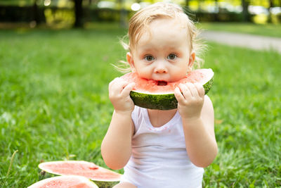 Funny smiling kid boy in white bodysuit eating watermelon at green lawn