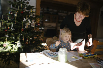 Grandmother looking at granddaughter painting on book in house during christmas