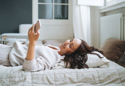 Young woman with brunette long hair in cozy knitted cardigan using mobile phone in bed at home