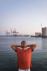 Young happy ethnic male traveler with raised arms standing behind rippled sea under sky in twilight