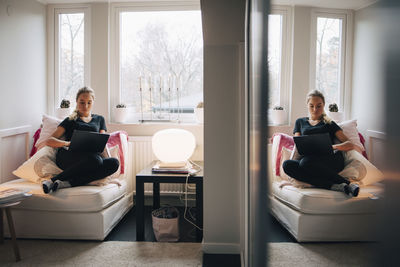 Reflection of teenage girl using laptop reclining on seat against window at home