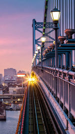 Illuminated train on track by bridge over river in city during sunset