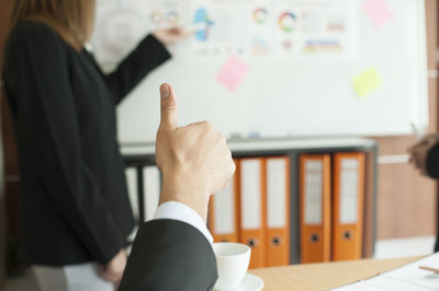 Cropped hand showing thumbs up sign with colleague giving presentation at office