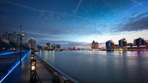 Illuminated buildings by river against sky at dusk