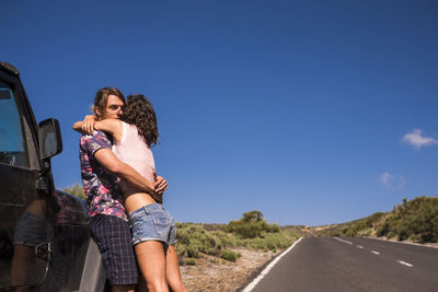 Young couple embracing on road against sky during sunny day