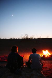 Rear view of woman and boy sitting by bonfire at sunset