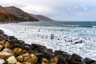 Scenic view of rocky beach in the coast of ireland against mountains and cloudy sky