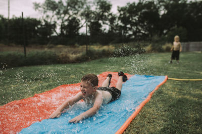 Boy sliding on water slide at yard with brother standing in background