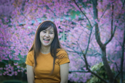 Portrait of smiling young woman with pink flower standing against trees