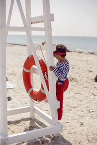 Child boy in striped clothes and red pants  standing on beach. white lifeguard tower, with a circle