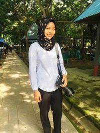 Portrait of smiling young woman with camera standing on footpath