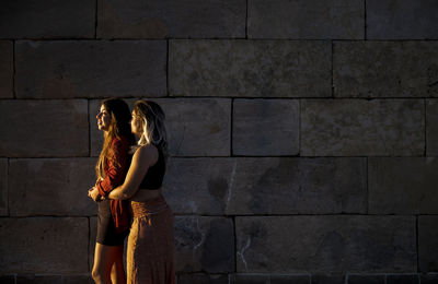 Two girls standing against stone wall during sunset