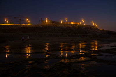 Illuminated city by sea against sky at night