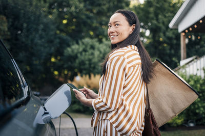 Smiling woman carrying bags and holding smart phone while charging car in back yard