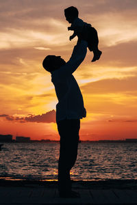 Silhouette man raising son while standing by sea against sky during sunset